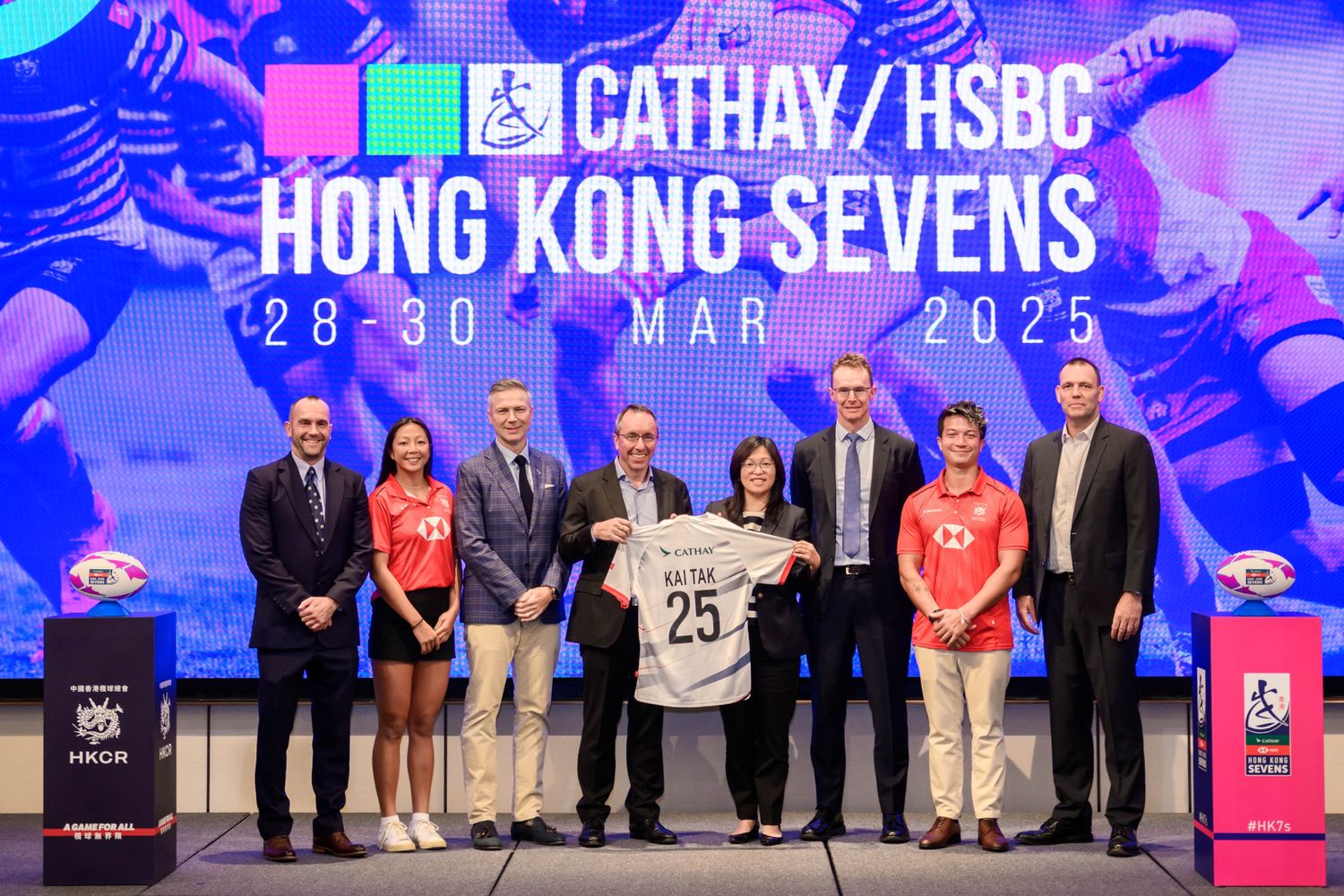 A group of individuals stands in front of a colorful backdrop promoting the Cathay/HSBC Hong Kong Sevens, holding a jersey with the name "KAI TAK" while showcasing rugby-related items and graphics.